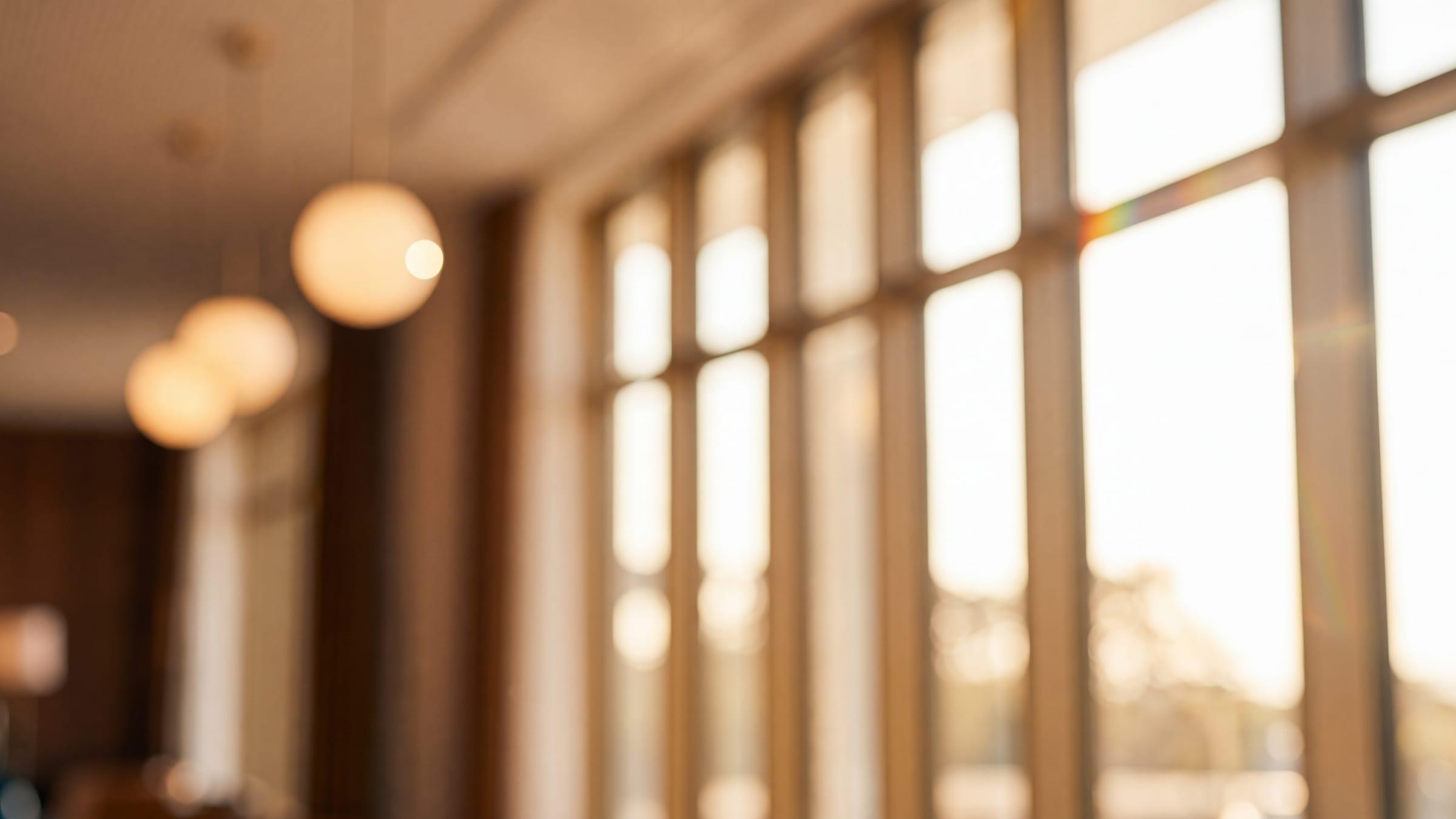 Lachender Hotelier im Profil in der Lobby seines Hotels, eine Mitarbeiterin steht unscharf im Hintergrund, Sonnenlicht fällt durch die Fenster.